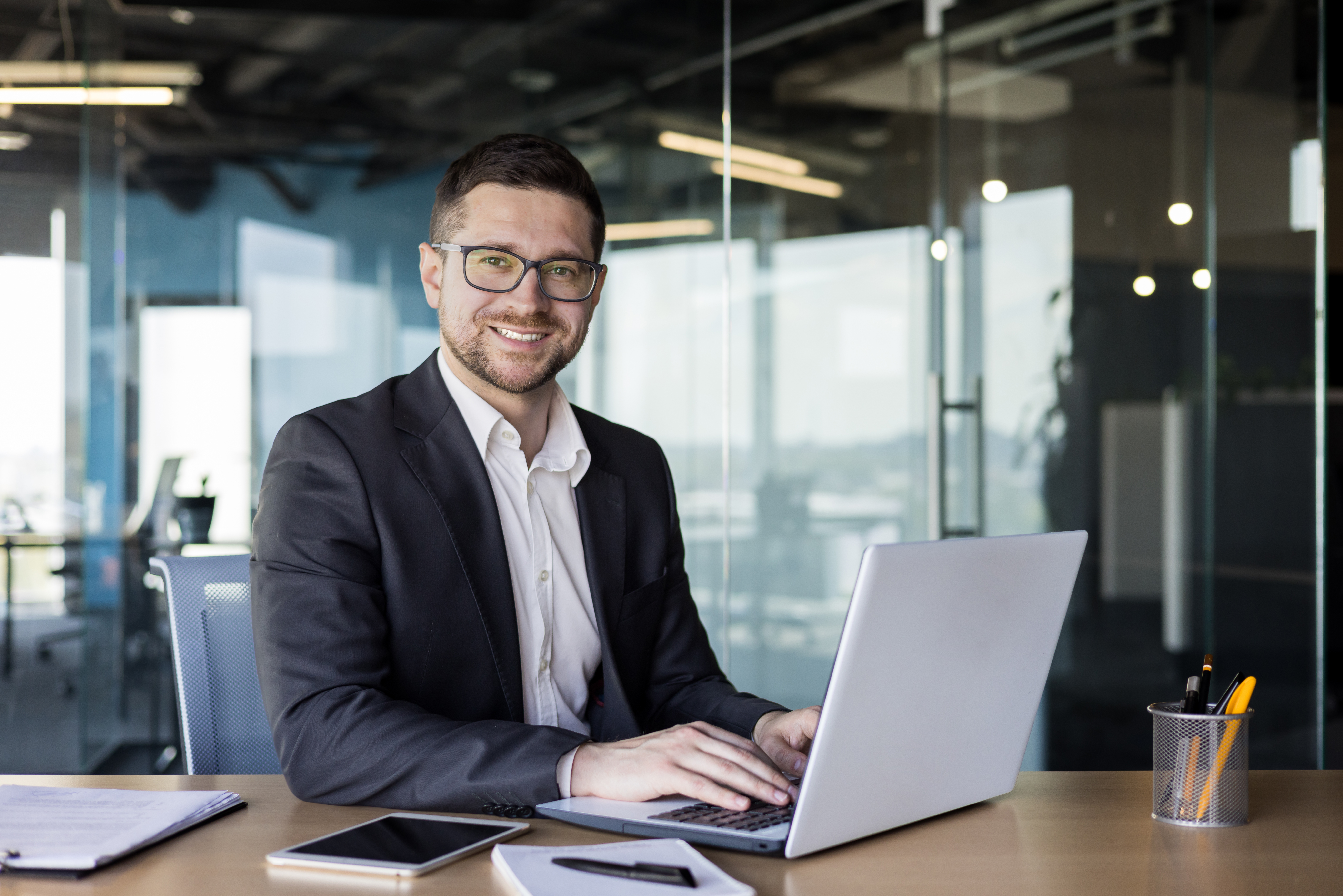 Portrait of a successful male businessman, banker, financial director sitting in the office at the table and using a laptop. Looking and smiling at the camera